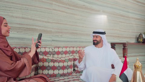 Documenting memorable moments via mobile, an Arab Gulf Emirati woman wearing an abaya and hijab taking a picture of her husband, gestures of pleasure and happiness, cafes and coffee shops in the Emirates, spending happy times