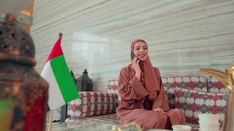 Documenting memorable moments, making a phone call using a mobile phone, checking on family and friends, an outing, gestures of pleasure and happiness, spending happy times, UAE cafes and coffee shops, an Emirati Gulf Arab woman sitting in a cafe with the UAE flag next to her