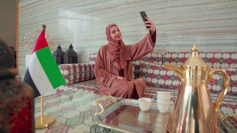 Checking on family and friends, going on an outing, making a phone call using a mobile phone, gestures of pleasure and happiness, spending happy times, UAE cafes and coffee shops, documenting memorable moments, an Emirati Gulf Arab woman sitting in a cafe with the UAE flag next to her