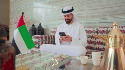 الدردشة عبر الانترنت ،تصفح مواقع التواصل الاجتماعي عبر الجوال ، رجل عربي خليجي اماراتي يرتدي الكندورة والغتره يجلس فى مقهي وبجانبه علم الامارات ، كافيهات ومقاهي الامارات،   نزهه فى الخارج ،ايماءات السرور والسعاده ، قضاء اوقات سعيده