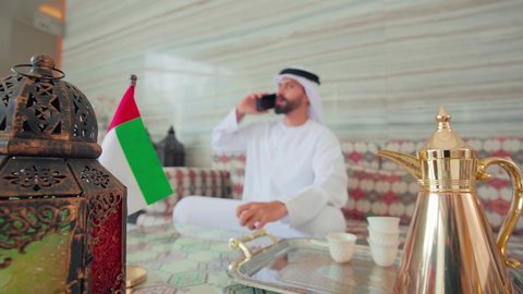الاطمئنان على الاهل والاصدقاء، اجراء مكالمه هاتفيه ،تصفح مواقع التواصل الاجتماعي عبر الجوال ، رجل عربي خليجي اماراتي يرتدي الكندورة والغتره يجلس فى مقهي وبجانبه علم الامارات ، كافيهات ومقاهي الامارات،   نزهه فى الخارج ،ايماءات السرور والسعاده ، قضاء اوقات سعيده