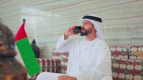 اجراء مكالمه هاتفيه ،تصفح مواقع التواصل الاجتماعي عبر الجوال ، رجل عربي خليجي اماراتي يرتدي الكندورة والغتره يجلس فى مقهي وبجانبه علم الامارات ، كافيهات ومقاهي الامارات،  الاطمئنان على الاهل والاصدقاء، نزهه فى الخارج ،ايماءات السرور والسعاده ، قضاء اوقات سعيده