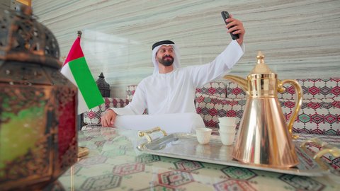 التقاط صور السيلفي ،تصفح مواقع التواصل الاجتماعي عبر الجوال ، رجل عربي خليجي اماراتي يرتدي الكندورة والغتره يجلس فى مقهي وبجانبه علم الامارات ، كافيهات ومقاهي الامارات،  الاطمئنان على الاهل والاصدقاء، نزهه فى الخارج ،ايماءات السرور والسعاده ، قضاء اوقات سعيده
