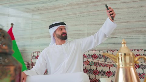 توثيق اللحظات التذكاريه ،تصفح مواقع التواصل الاجتماعي عبر الجوال ، رجل عربي خليجي اماراتي يرتدي الكندورة والغتره يجلس فى مقهي وبجانبه علم الامارات ، كافيهات ومقاهي الامارات،  الاطمئنان على الاهل والاصدقاء، نزهه فى الخارج ،ايماءات السرور والسعاده ، قضاء اوقات سعيده