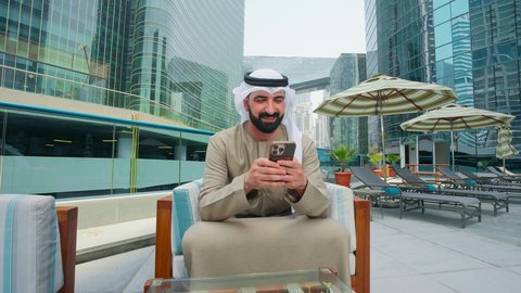 قضاء اوقات ممتعه فى الخارج، تصفح مواقع التواصل الاجتماعي ، استنشاق الهواء النقي ، رجل عربي خليجي اماراتي يرتدي الكندورة والغتره يجلس على كرسي حول المسبح في أحد الفنادق في المملكة العربية السعودية يستخدم الهاتف المحمول ،  القيام بالأنشطة الترفيهية المسلية,استخدام التقنيات الحديثة ،شاليهات و منتجعات فاخرة