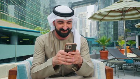 Browsing social media, breathing fresh air, having a good time outside, an Emirati Gulf Arab man wearing a kandura and ghutra sitting on a chair around the pool in a hotel in the Kingdom of Saudi Arabia using a mobile phone, doing fun recreational activities, using modern technology, chalets and luxury resorts
