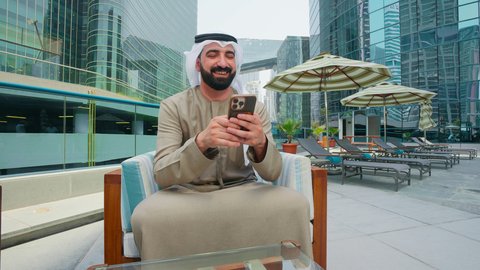 التقاط صور السيلفي ، تصفح مواقع التواصل الاجتماعي ، استنشاق الهواء النقي ، قضاء اوقات ممتعه فى الخارج، رجل عربي خليجي اماراتي يرتدي الكندورة والغتره يجلس على كرسي حول المسبح في أحد الفنادق في المملكة العربية السعودية يستخدم الهاتف المحمول ،  القيام بالأنشطة الترفيهية المسلية,استخدام التقنيات الحديثة ،شاليهات و منتجعات فاخرة