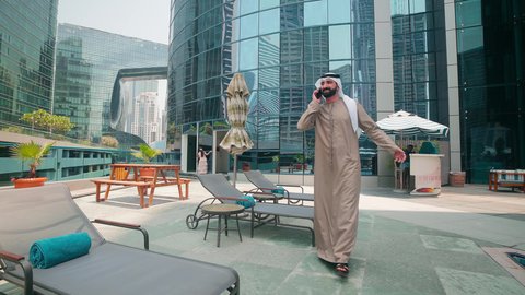 Breathing fresh air, having a good time outside, an Emirati Gulf Arab man wearing a kandura and ghutra walking around the pool in a hotel in Saudi Arabia using a mobile phone, doing fun recreational activities, using modern technology, luxury chalets and resorts, making a phone call