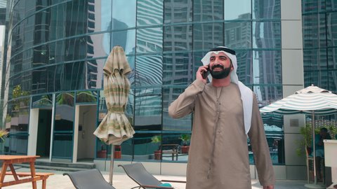 Having a good time outside, an Emirati Gulf Arab man wearing a kandura and ghutra walking around the pool in a hotel in Saudi Arabia using a mobile phone, breathing fresh air, doing fun recreational activities, using modern technology, luxury chalets and resorts, making a phone call