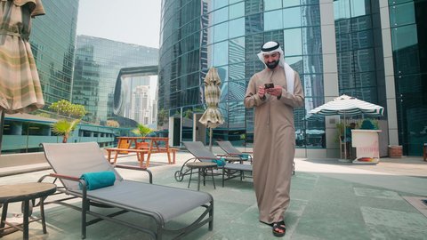 An Emirati Gulf Arab man wearing a kandura and ghutra walking around the pool in a hotel in Saudi Arabia using a mobile phone, breathing fresh air, doing fun recreational activities, using modern technology, luxury chalets and resorts, making a phone call