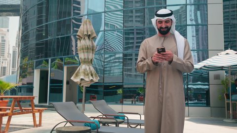 استنشاق الهواء النقي ، القيام بالأنشطة الترفيهية المسلية,استخدام التقنيات الحديثة ،شاليهات و منتجعات فاخرة ، رجل عربي خليجي اماراتي يرتدي الكندورة والغتره يتجول حول المسبح في أحد الفنادق في المملكة العربية السعودية يستخدم الهاتف المحمول ،اجراء مكالمة هاتفية