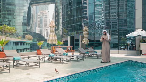 استخدام التقنيات الحديثة ،شاليهات و منتجعات فاخرة ، رجل عربي خليجي اماراتي يرتدي الكندورة والغتره يتجول حول المسبح في أحد الفنادق في المملكة العربية السعودية يستخدم الهاتف المحمول ،اجراء مكالمة هاتفية ، القيام بالأنشطة الترفيهية المسلية, استنشاق الهواء النقي
