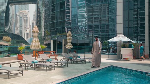 شاليهات و منتجعات فاخرة ، رجل عربي خليجي اماراتي يرتدي الكندورة والغتره يتجول حول المسبح في أحد الفنادق في المملكة العربية السعودية يستخدم الهاتف المحمول ، استخدام التقنيات الحديثة ،اجراء مكالمة هاتفية ، القيام بالأنشطة الترفيهية المسلية, استنشاق الهواء النقي