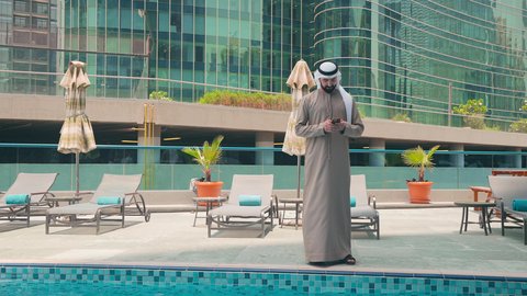 اجراء مكالمة هاتفية ، رجل عربي خليجي اماراتي يرتدي الكندورة والغتره يتجول حول المسبح في أحد الفنادق في المملكة العربية السعودية يستخدم الهاتف المحمول ،شاليهات و منتجعات فاخرة ،استخدام التقنيات الحديثة ، القيام بالأنشطة الترفيهية المسلية,  استنشاق الهواء النقي