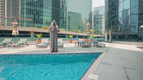 رجل عربي خليجي اماراتي يرتدي الكندورة والغتره يتجول حول المسبح في أحد الفنادق في المملكة العربية السعودية يستخدم الهاتف المحمول ،شاليهات و منتجعات فاخرة ،  استخدام التقنيات الحديثة ،اجراء مكالمة هاتفية ، القيام بالأنشطة الترفيهية المسلية, استنشاق الهواء النقي