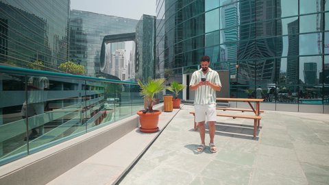 استخدام التقنيات الحديثة، مشاهدة التطور العمراني في المدينة، التجول واستنشاق الهواء النقي في الخارج,  الفخر والاعتزاز بالتقدم المعماري ،  الدردشة عبر الانترنت ، النمو العمراني في الامارات ،رجل عربي خليجي اماراتي يرتدي الزي الكاجوال يستخدم الهاتف المحمول