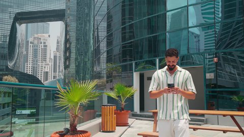 مشاهدة التطور العمراني في المدينة، التجول واستنشاق الهواء النقي في الخارج,  الفخر والاعتزاز بالتقدم المعماري ،  الدردشة عبر الانترنت ، النمو العمراني في الامارات ،رجل عربي خليجي اماراتي يرتدي الزي الكاجوال يستخدم الهاتف المحمول