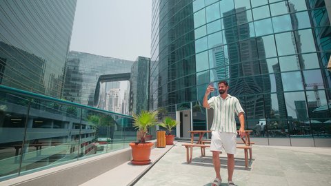 Walking around and breathing fresh air outside, being proud of architectural progress, chatting online, urban growth in the Emirates, watching the urban development in the city, an Arab Gulf Emirati man wearing casual clothes using a mobile phone