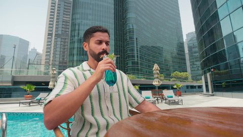 Luxury chalets and resorts, doing fun recreational activities, an Emirati Gulf Arab man wearing casual clothes enjoying sitting at a table around the pool in a hotel in the Kingdom of Saudi Arabia, spending happy times, enjoying refreshing summer drinks