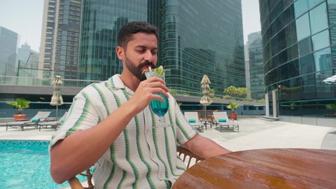 An Arab Gulf Emirati man wearing casual clothes enjoying sitting at a table around the pool in one of the hotels in the Kingdom of Saudi Arabia, luxury chalets and resorts, spending happy times, doing entertaining recreational activities, and enjoying refreshing summer drinks