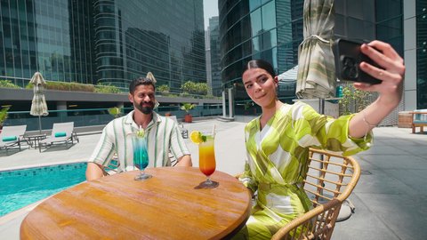 التقاط صور السيلفي باستخدام الجوال ،توثيق اللحظات التذكارية ، شاليهات و منتجعات فاخرة،  الاستمتاع بالمشروبات الصيفية المنعشة, زوجان عربيان خليجيان اماراتيين يرتديان الزي الكاجوال يستمتعان بالحديث على طاولة حول المسبح في أحد الفنادق في المملكة العربية السعودية