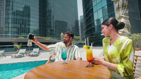 استخدام التقنيات الحديثة ، التقاط صور السيلفي باستخدام الجوال ،توثيق اللحظات التذكارية ،  الاستمتاع بالمشروبات الصيفية المنعشة, زوجان عربيان خليجيان اماراتيين يرتديان الزي الكاجوال يستمتعان بالحديث على طاولة حول المسبح في أحد الفنادق في المملكة العربية السعودية
