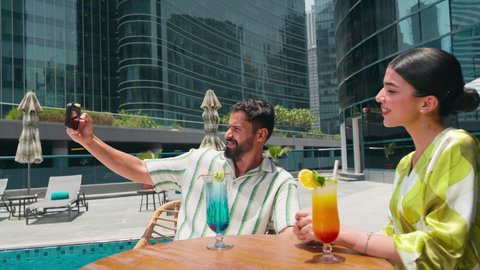 توثيق اللحظات التذكارية ،التقاط صور السيلفي باستخدام الجوال ، شاليهات و منتجعات فاخرة،  الاستمتاع بالمشروبات الصيفية المنعشة, زوجان عربيان خليجيان اماراتيين يرتديان الزي الكاجوال يستمتعان بالحديث على طاولة حول المسبح في أحد الفنادق في المملكة العربية السعودية