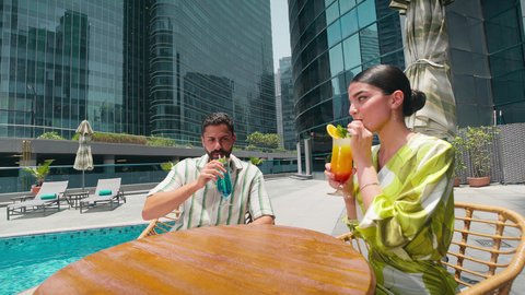 الاستمتاع بالمشروبات الصيفية المنعشة, القيام بالأنشطة الترفيهية المسلية, زوجان عربيان خليجيان اماراتيين يرتديان الزي الكاجوال يستمتعان بالحديث على طاولة حول المسبح في أحد الفنادق في المملكة العربية السعودية، شاليهات و منتجعات فاخرة