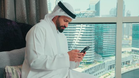 تصفح موقع الانترنت، الدردشة مع الأصدقاء والأقارب، إرسال رسالة نصية عبر الجوال, تصوير من الجانب لرجل عربي خليجي اماراتي يرتدي الكندورة و الغتره يجلس على كرسي بجانب النافذة فى المنزل يقوم بمتابعة آخر أخبار السوشال ميديا عبر الهاتف المحمول ، دمج التكنولوجيا الحديثة بالحياة العملية، النظر الى شيء ما