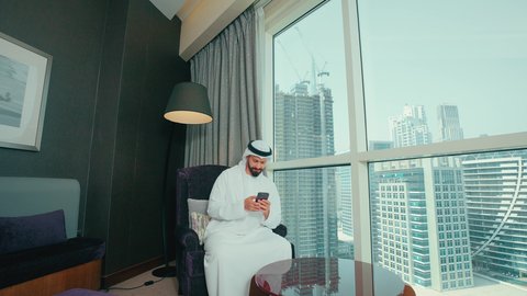 الدردشة مع الأصدقاء والأقارب، إرسال رسالة نصية عبر الجوال, رجل عربي خليجي اماراتي يرتدي الكندورة و الغتره يجلس على كرسي بجانب النافذة فى المنزل يقوم بمتابعة آخر أخبار السوشال ميديا عبر الهاتف المحمول ، تصفح موقع الانترنت، دمج التكنولوجيا الحديثة بالحياة العملية، النظر الى شيء ما