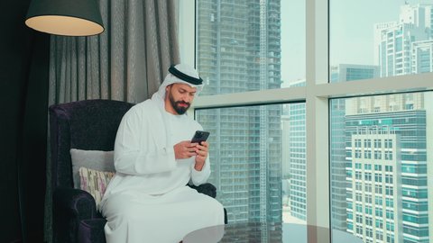 إرسال رسالة نصية عبر الجوال, رجل عربي خليجي اماراتي يرتدي الكندورة و الغتره يجلس على كرسي بجانب النافذة فى المنزل يقوم بمتابعة آخر أخبار السوشال ميديا عبر الهاتف المحمول, التواصل مع الأصدقاء والأقارب ، تصفح موقع الانترنت، دمج التكنولوجيا الحديثة بالحياة العملية، النظر الى شيء ما