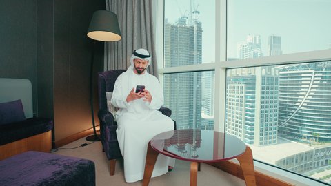 ايماءات السعادة و السرورة ، توثيق اللحظات السعيدة والمميزة, استخدام الاجهزة والتقنيات الحديثة, رجل عربي خليجي اماراتي  مبتسم يرتدي الكندورة و الغتره يجلس على كرسي بجانب النافذة فى المنزل يقوم بالتقاط صور السيلفي عبر الهاتف المحمول