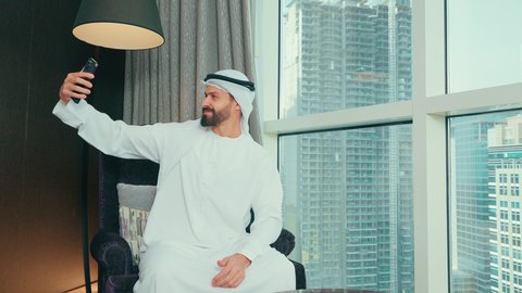 توثيق اللحظات السعيدة والمميزة, استخدام الاجهزة والتقنيات الحديثة, رجل عربي خليجي اماراتي  مبتسم يرتدي الكندورة و الغتره يجلس على كرسي بجانب النافذة فى المنزل يقوم بالتقاط صور السيلفي عبر الهاتف المحمول بإيماءات السرور