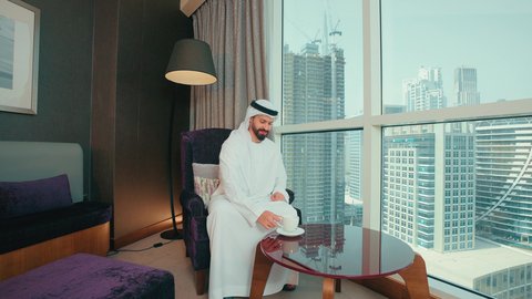 تناول الأعشاب الشتوية الدافئة،  قضاء وقت الفراغ باحتساء المشروب المفضل، الاستمتاع بالمشروبات الساخنة، رجل عربي خليجي اماراتي يرتدي الكندورة والغترة البيضاء يجلس على كرسي بجانب النافذة فى المنزل يقوم بشرب القهوة، إيماءات السعادة والسرور، النظر الى شيء ما