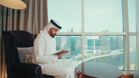 ايماءات الاندماج و التركيز، قضاء وقت الفراغ بالقراءة والتعلم والثقافة، مفهوم التعلم الذاتي، مطالعة القصص الممتعة،رجل عربي خليجي اماراتي يرتدي الكندورة و الغتره يجلس بجانب النافذة على كرسي مريح يقوم بمطالعة كتاب، التزود بالمعلومات من الكتب والروايات