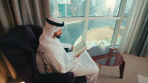 مطالعة القصص الممتعة، مفهوم التعلم الذاتي، قضاء وقت الفراغ بالقراءة والتعلم والثقافة، تصوير من الجانب لرجل عربي خليجي اماراتي يرتدي الكندورة و الغتره يجلس بجانب النافذة على كرسي مريح يقوم بمطالعة كتاب، التزود بالمعلومات من الكتب والروايات، ايماءات الاندماج و التركيز