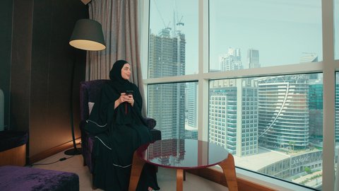 Communication and chatting with family and friends, browsing social media sites, spending enjoyable times through social media applications, an Arab Gulf Emirati woman wearing an abaya and hijab sitting on a chair by the window using a mobile phone, looking at something.