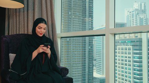 التواصل والدردشة مع الأهل والأصدقاء, تصفح مواقع التواصل الاجتماعي, قضاء أوقات ممتعه عبر تطبيقات السوشال ميديا, امراة عربيه خليجيه اماراتية ترتدي العباءة و الحجاب تجلس على كرسي بجانب النافذة تقوم باستخدام الهاتف المحمول، النظر الى شيء ما