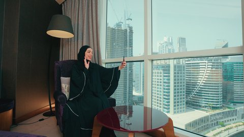 اجراء مكالمة فيديو ، رفع كف الايد بتعابير التحية و السلام، التواصل مع العائلة والأصدقاء، مشاركة الأخبار والأحاديث,سيده سعودية تستخدم الأجهزة التقنية الحديثة في الحياة اليومية, امراة عربية خليجية اماراتية محجبة ترتدي العباءة تجلس على كرسي بجانب النافذة تقوم بإجراء اتصال عبر الهاتف المحمول، ايماءات السعادة و السرور