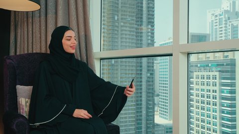 اجراء مكالمة فيديو ، رفع كف الايد بتعابير التحية و السلام، التواصل مع العائلة والأصدقاء، مشاركة الأخبار والأحاديث,سيده سعودية تستخدم الأجهزة التقنية الحديثة في الحياة اليومية, امراة عربية خليجية اماراتية محجبة ترتدي العباءة تجلس على كرسي بجانب النافذة تقوم بإجراء اتصال عبر الهاتف المحمول، ايماءات السعادة و السرور