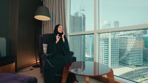 مشاركة الأخبار والأحاديث, التواصل مع العائلة والأصدقاء, استخدام الأجهزة التقنية الحديثة في الحياة اليومية, امراة عربية خليجية اماراتية محجبة ترتدي العباءة تجلس على كرسي بجانب النافذة تقوم بإجراء اتصال هاتفي عبر الهاتف المحمول، ايماءات السعادة و السرور