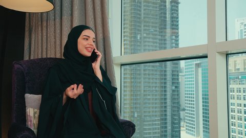 Sharing news and conversations, communicating with family and friends, using modern technological devices in daily life, an Arab Gulf Emirati woman wearing a hijab and an abaya sitting on a chair by the window making a phone call on her mobile phone, gestures of happiness and joy.