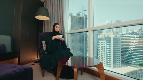 Warm and calm atmosphere, gestures of happiness and joy, a Saudi woman looking at something, drinking a hot and delicious beverage, sitting and enjoying hot drinks and warm winter herbs, an Emirati Gulf Arab woman wearing a hijab and an abaya sitting on a chair by the window in the house.