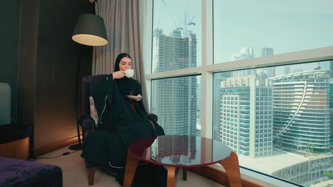 ايماءات السعادة و السرور، سيده سعودية تنظر الى شيء ما، شرب مشروب ساخن ولذيذ، الجلوس والاستمتاع بالمشروبات الساخنة والأعشاب الشتوية الدافئة,  امرأة عربية خليجية اماراتية محجبة ترتدي عباءة تجلس على كرسي بجانب النافذة فى المنزل