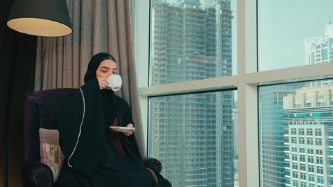 سيده سعودية تنظر الى شيء ما، شرب مشروب ساخن ولذيذ، الجلوس والاستمتاع بالمشروبات الساخنة والأعشاب الشتوية الدافئة,  امرأة عربية خليجية اماراتية محجبة ترتدي عباءة تجلس على كرسي بجانب النافذة فى المنزل ، ايماءات السعادة و السرور