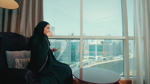 شرب مشروب ساخن ولذيذ، الجلوس والاستمتاع بالمشروبات الساخنة والأعشاب الشتوية الدافئة, تصوير من الجانب امرأة عربية خليجية اماراتية محجبة ترتدي عباءة تجلس على كرسي بجانب النافذة فى المنزل ، ايماءات السعادة و السرور ،سيده سعودية تنظر الى شيء ما