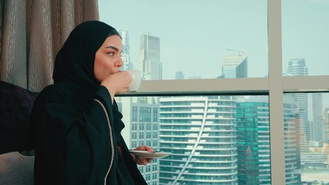 الجلوس والاستمتاع بالمشروبات الساخنة والأعشاب الشتوية الدافئة, شرب مشروب ساخن ولذيذ, تصوير من الجانب امرأة عربية خليجية اماراتية محجبة ترتدي عباءة تجلس على كرسي بجانب النافذة فى المنزل ، ايماءات السعادة و السرور ،سيده سعودية تنظر الى شيء ما