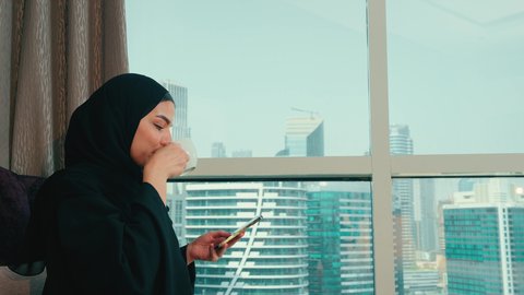 تعابير السرور والاندماج، تصفح التطبيقات وبرامج التواصل الاجتماعي، تبادل الأحاديث والأخبار مع الأهل والأصدقاء،تصوير من الجانب لامراة عربية خليجية اماراتية ترتدي العباءة والحجاب تمسك بيدها الهاتف المحمول تجلس بجانب النافذة فى المنزل، استخدام جهاز تقني حديث ومتطور، سيده سعودية تنظر الى شيء ما ، الاستمتاع بتناول المشروبات