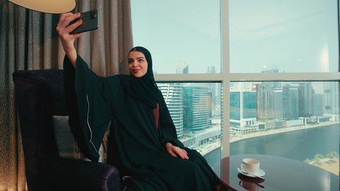 إجراء مكالمة فيديو عبر الجوال، توثيق اللحظات السعيدة, امراة عربية خليجية اماراتية ترتدي عباءة و الحجاب تقف بجانب النافذة فى المنزل تقوم بإلتقاط صورة سيلفي تذكارية بالهاتف المحمول بإيماءات تدل على السعادة ، سيده سعودية تقوم بإجراء مكالمة فيديو