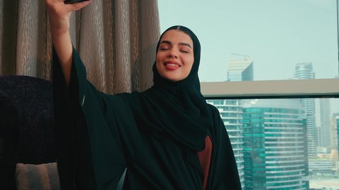 Documenting happy moments, an Arab Gulf Emirati woman wearing an abaya and hijab stands by the window in the house taking a commemorative selfie with her mobile phone with gestures indicating happiness, a Saudi woman is making a video call.
